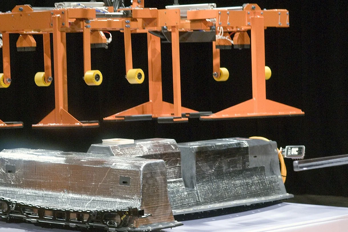 An orange handling arm with yellow castors above a grey T-shaped battery pack on a black background