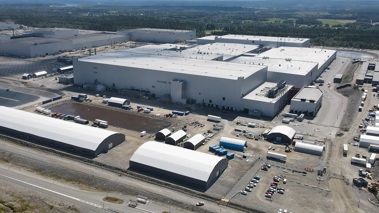 Aerial view of a large factory with white roofs surrounded by car parks, rounded sheds and forest, under a clear sky