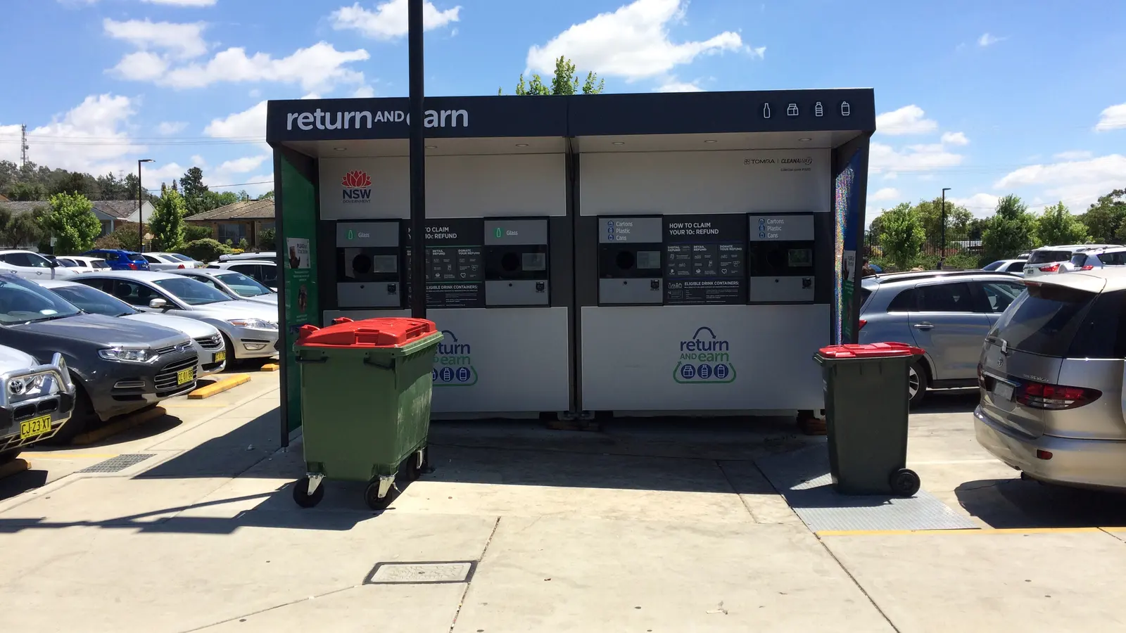 Deposit return machines for bottles and cans under a dark canopy, in a shopping centre car park, with 2 green bins