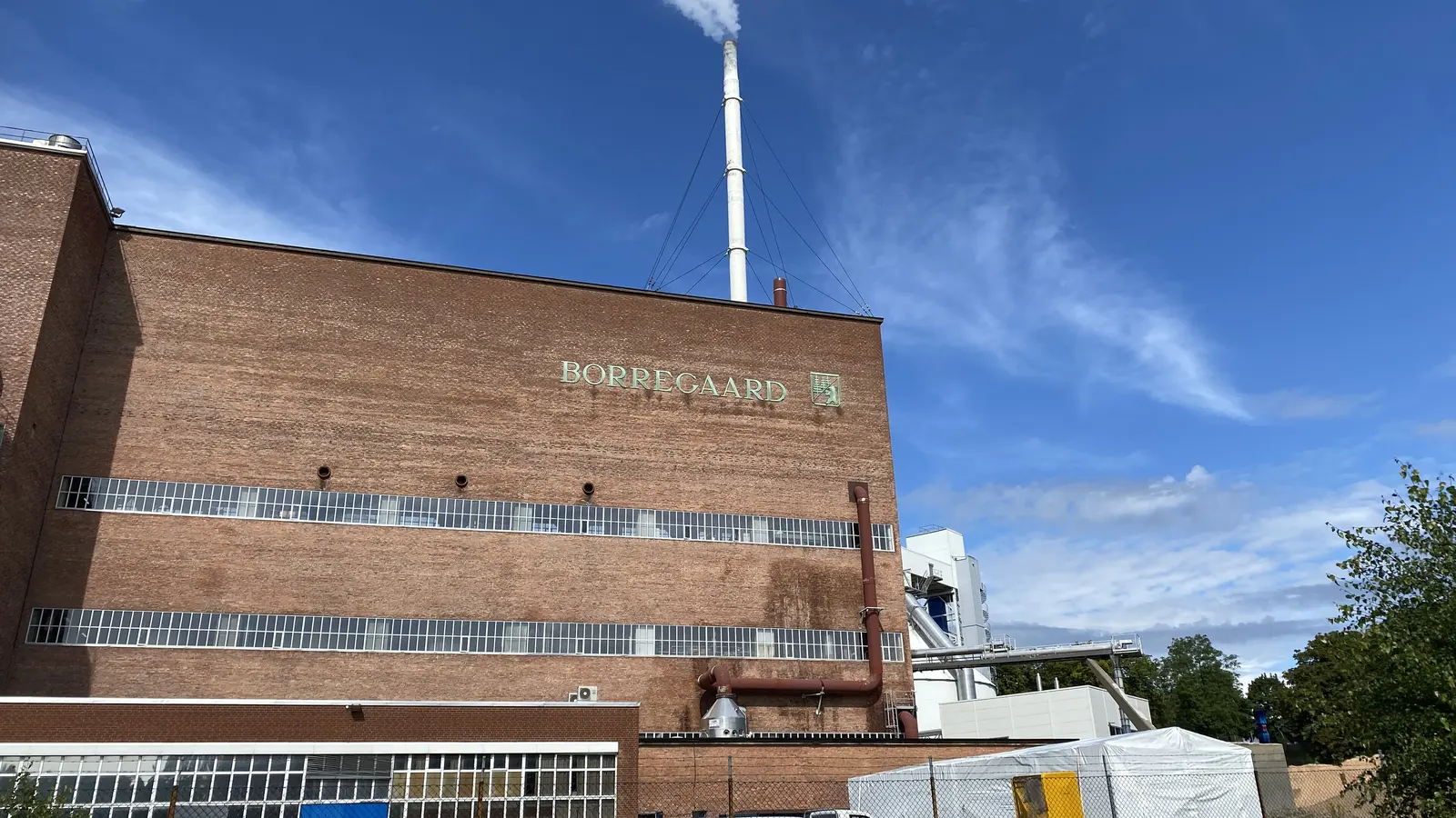 A red-brick industrial building bearing the name Borregaard, with a tall smoking chimney under a blue sky