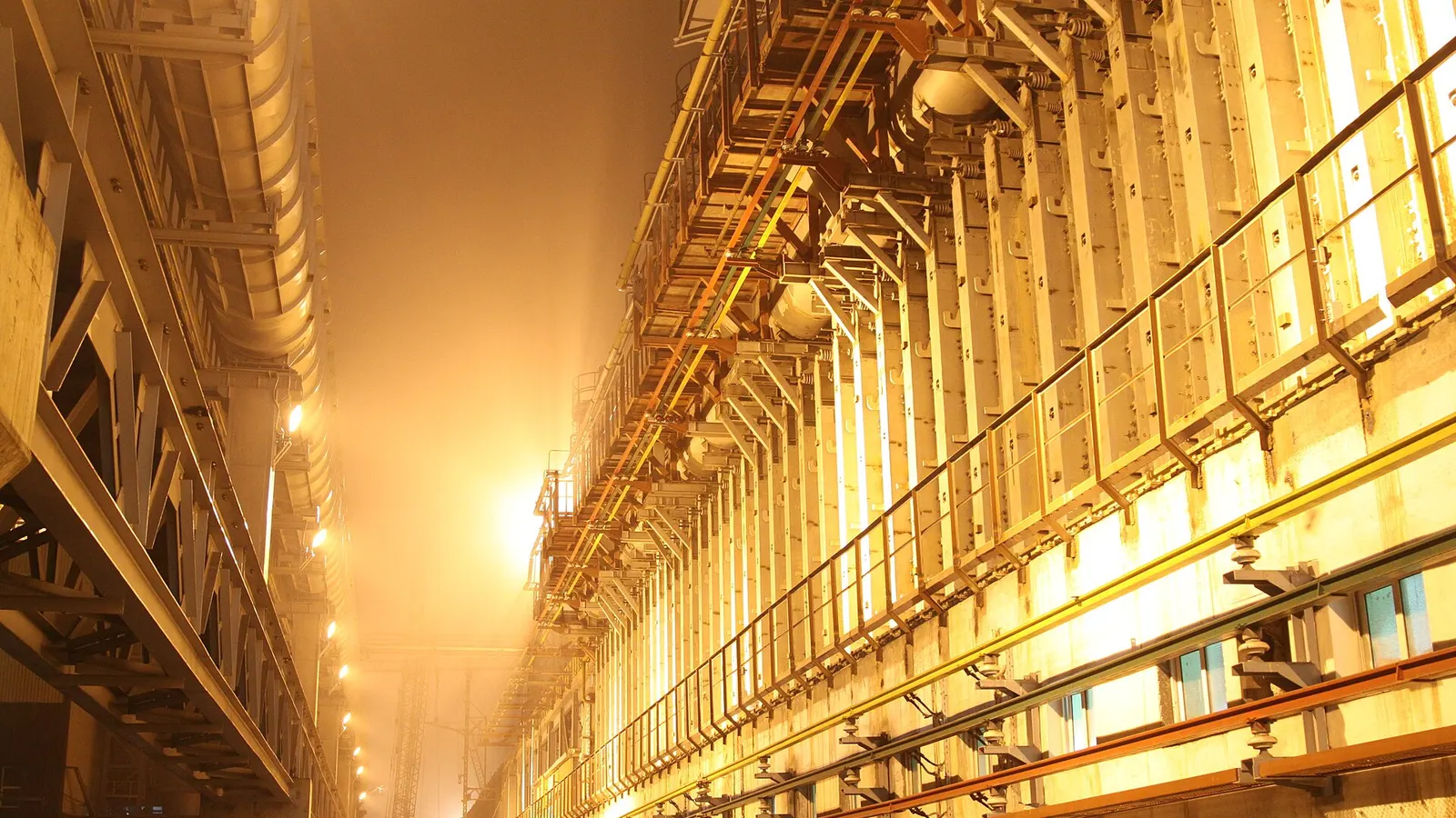 The facade of a coke oven battery lit in orange-yellow, with its walkways, rails and doors lined up, in a golden steam