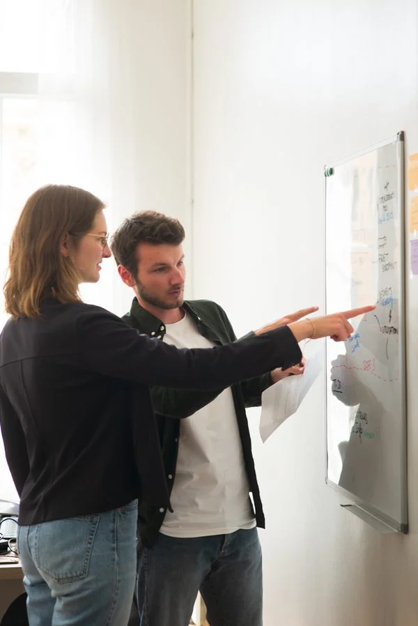 Two Projet Celsius consultants in front of a whiteboard, one pointing at a diagram while the other holds sheets of paper