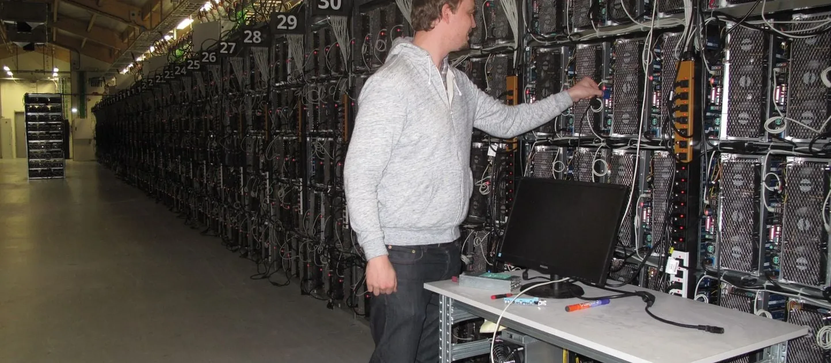 Bitcoin mining farm: an aisle of ASIC machines in rows, with a technician in a grey jumper