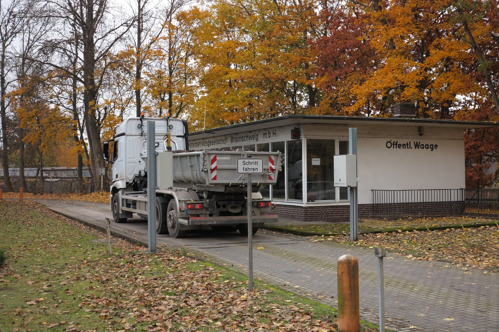 A rigid tipper truck stopped on a weighbridge in front of the glazed weighing booth, under orange autumn trees