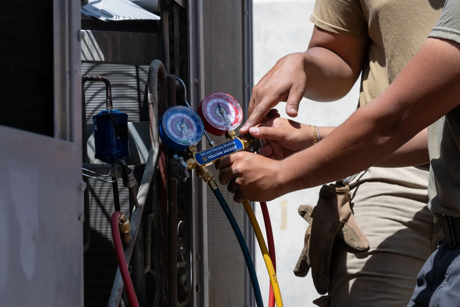 Two hands adjust a blue and red gauge manifold connected to the outdoor unit of an air conditioner, with blue, yellow and red hoses