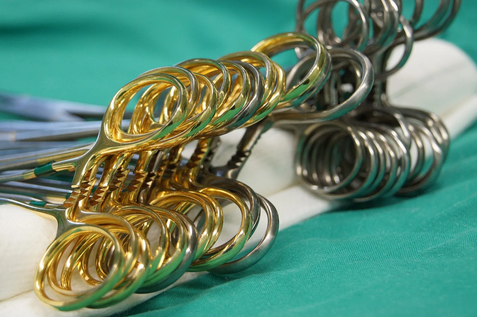 Steel surgical scissors and forceps, some with gold-coloured rings, lying on a green drape in an operating theatre