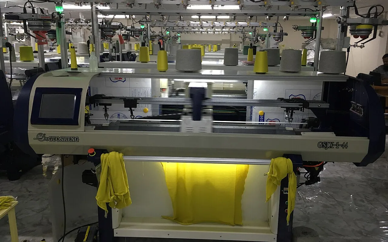 Computerised flat knitting machine, cones of yellow yarn above and a yellow jumper being knitted, in front of other machines in a row
