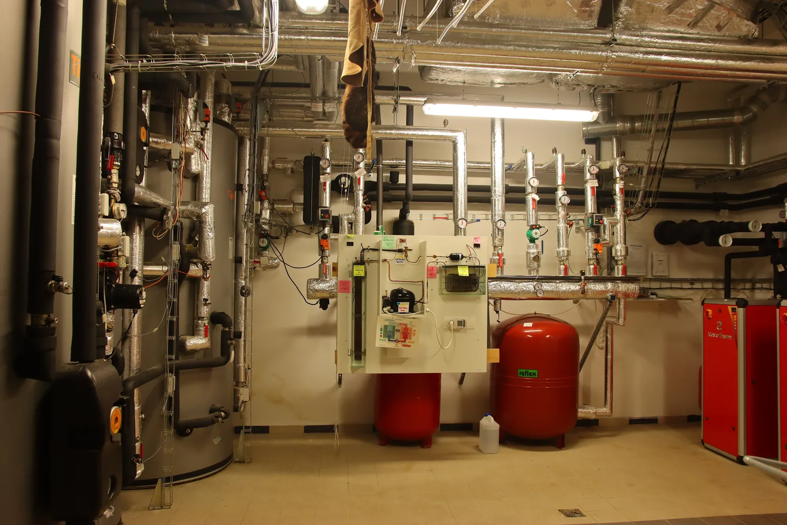 Bright boiler room: a large grey tank, insulated pipework, 2 red expansion vessels and, on the right, a red boiler