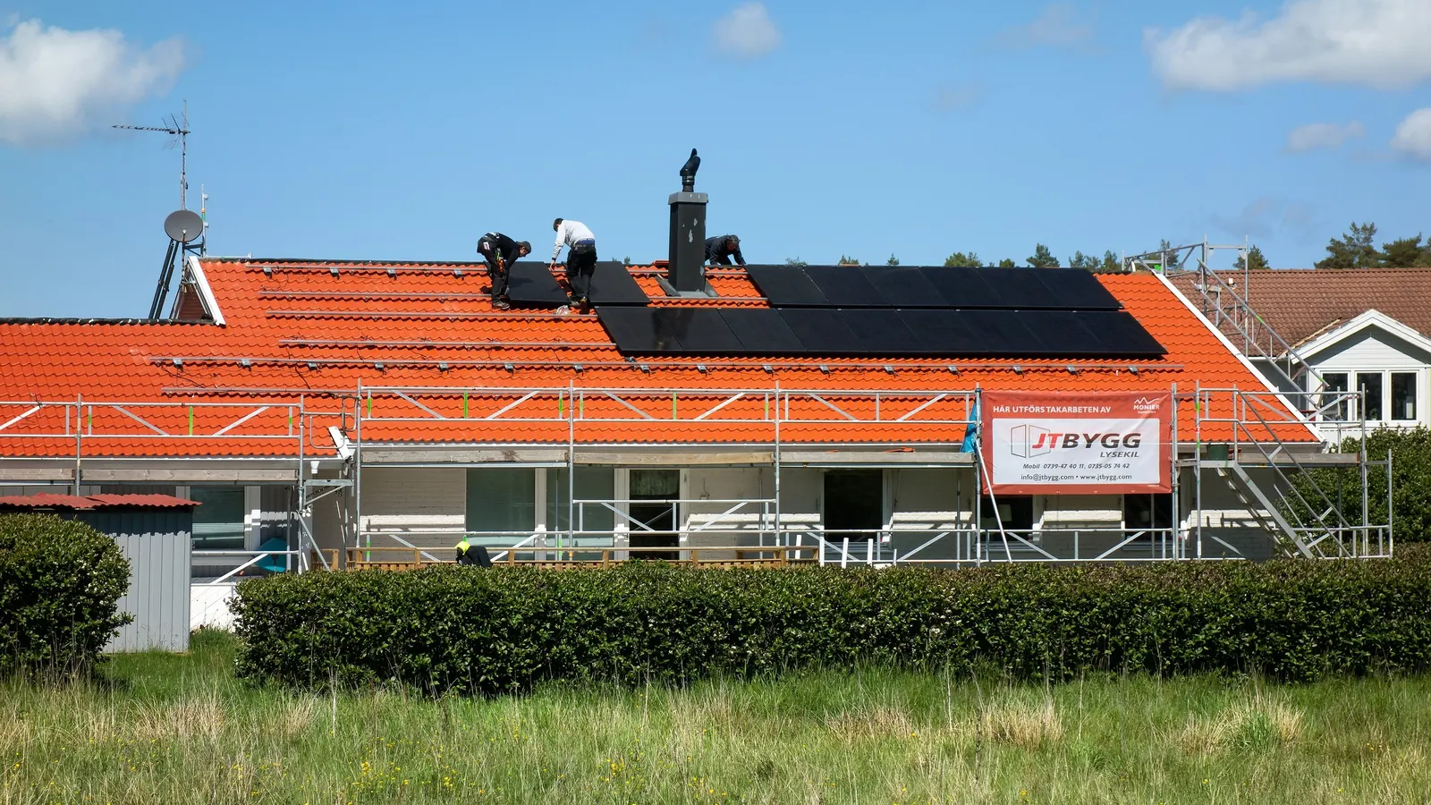 3 installers fitting black solar panels on the orange tiled roof of a house, under a blue sky, with scaffolding against the facade