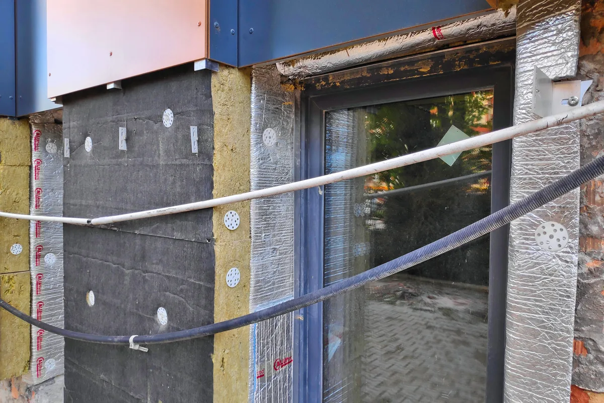 Facade under thermal renovation: mineral wool boards fixed with dowels, thin aluminium insulation around a glazed door, under blue cladding