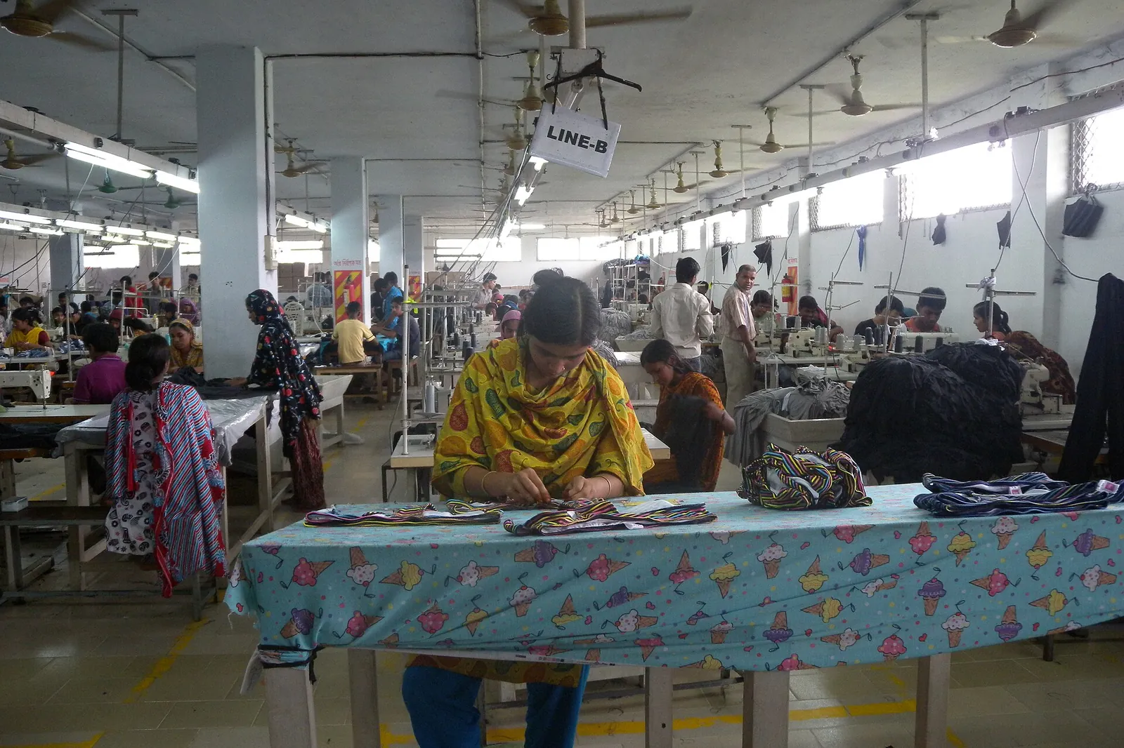 Garment factory in Bangladesh: women workers assembling clothes at long tables under ceiling fans
