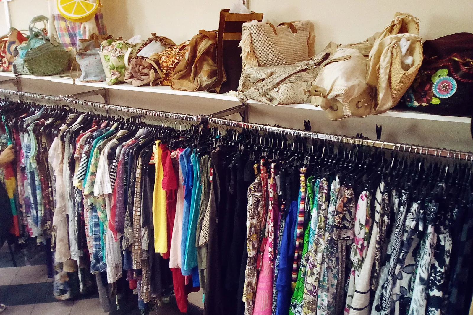 Rails of colourful used clothing in a second-hand shop, beneath a shelf of bags