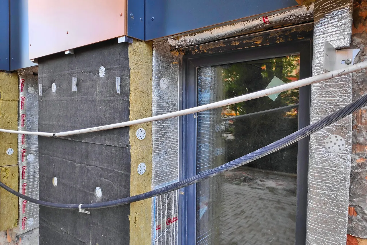 Facade under external insulation: yellow rock wool panels and black panels fixed with white dowels, thin silver insulation around a glazed door, metal cladding cassettes above