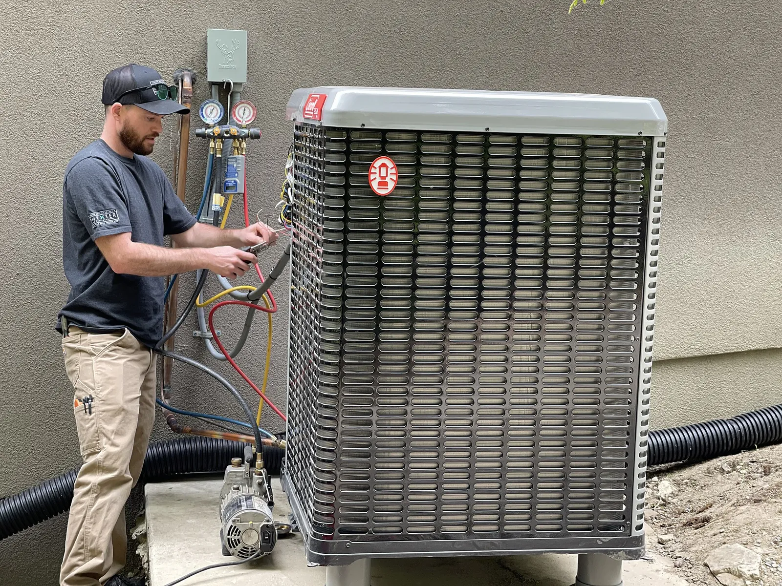 Refrigeration technician connecting a pressure-gauge manifold to the outdoor unit of a heat pump, with a vacuum pump on the ground