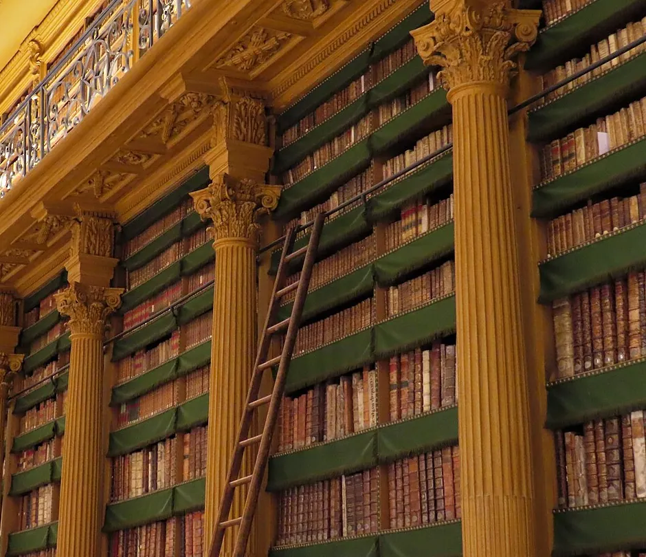 Shelves of old books running the full height of a room, with a ladder and a wrought-iron gallery