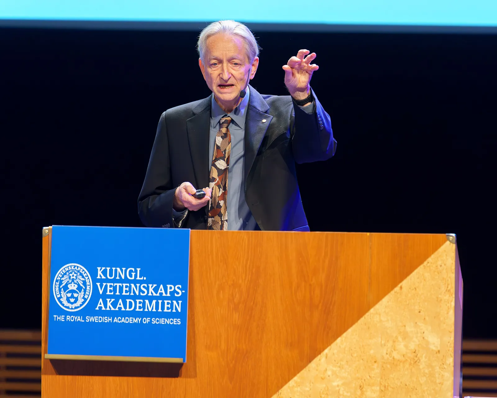 Geoffrey Hinton standing at a lectern, hand raised, during his Nobel lecture in Stockholm