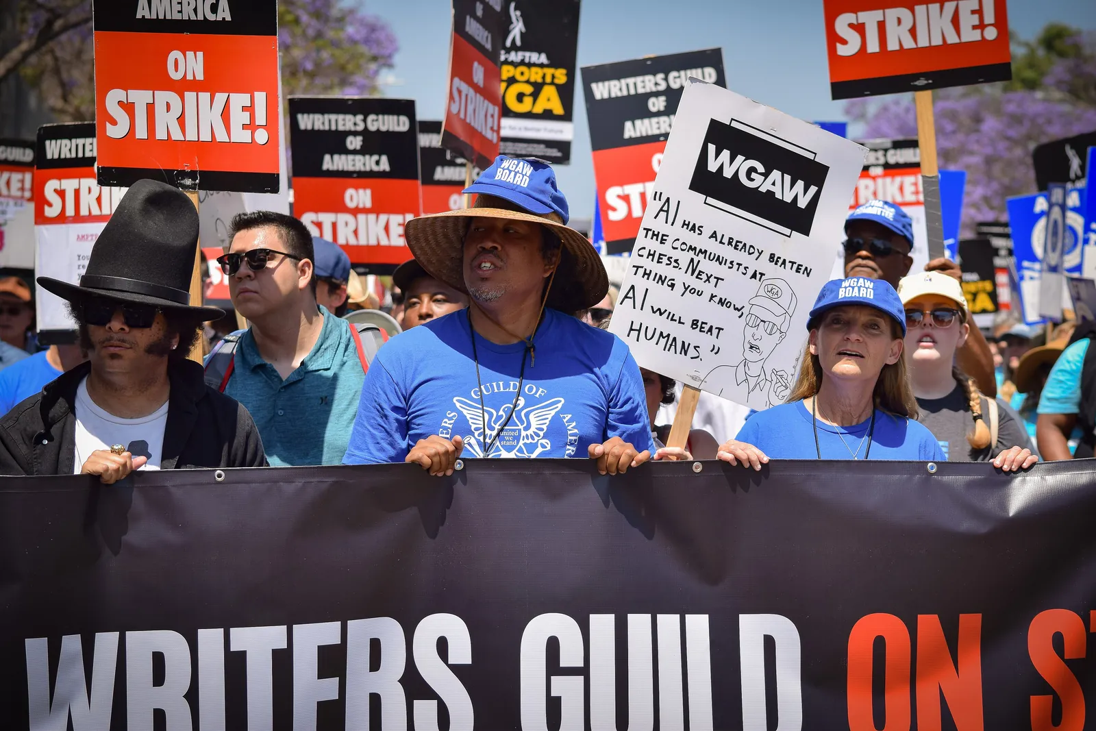 A march of striking screenwriters behind a black Writers Guild banner, red signs held up