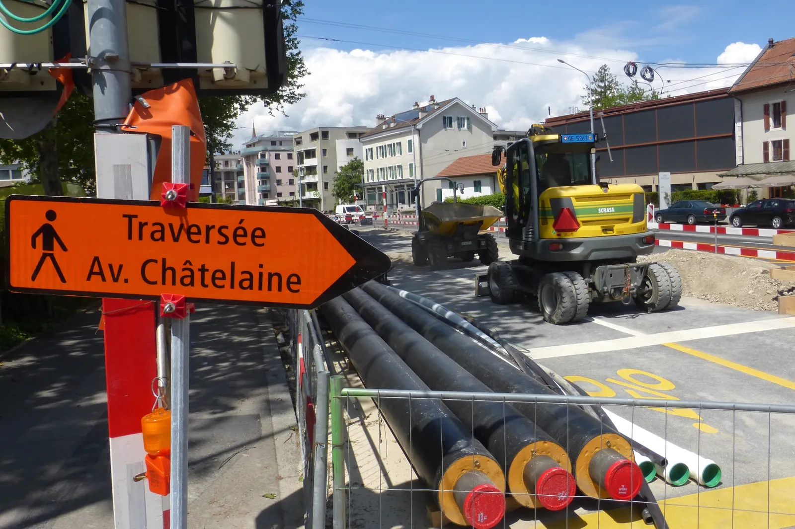 Black pipes with yellow insulation stored behind a construction-site fence, with an excavator and buildings in the background