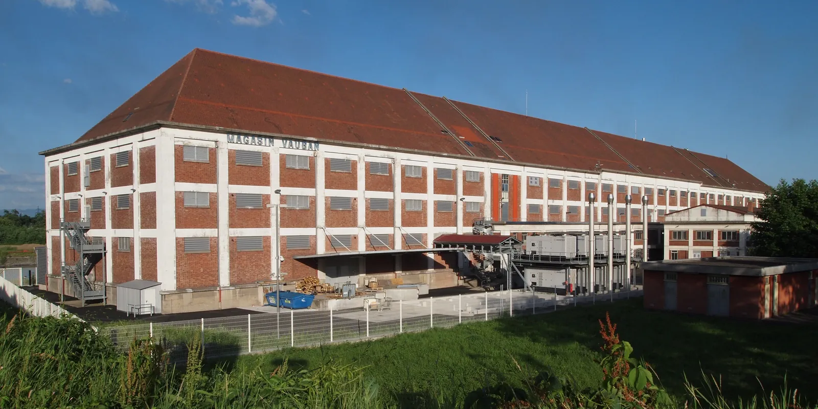 A former brick port warehouse bearing the inscription Magasin Vauban, flanked by technical units and exhaust stacks