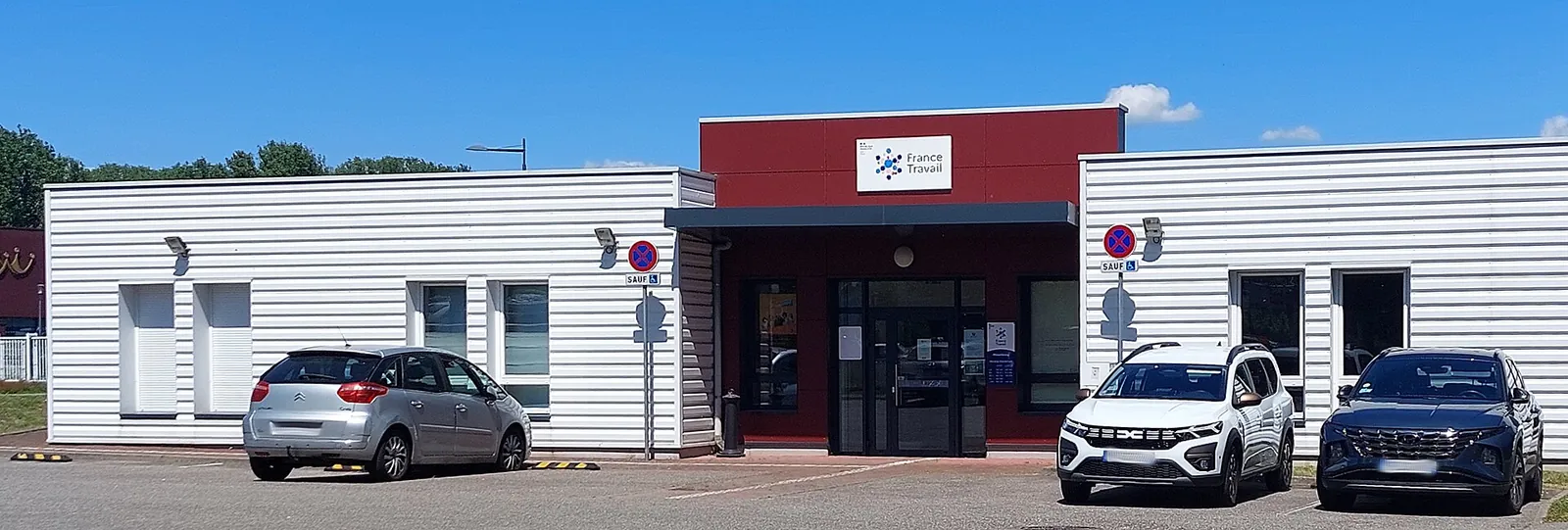 The white and red façade of a France Travail agency, seen from its car park under a blue sky