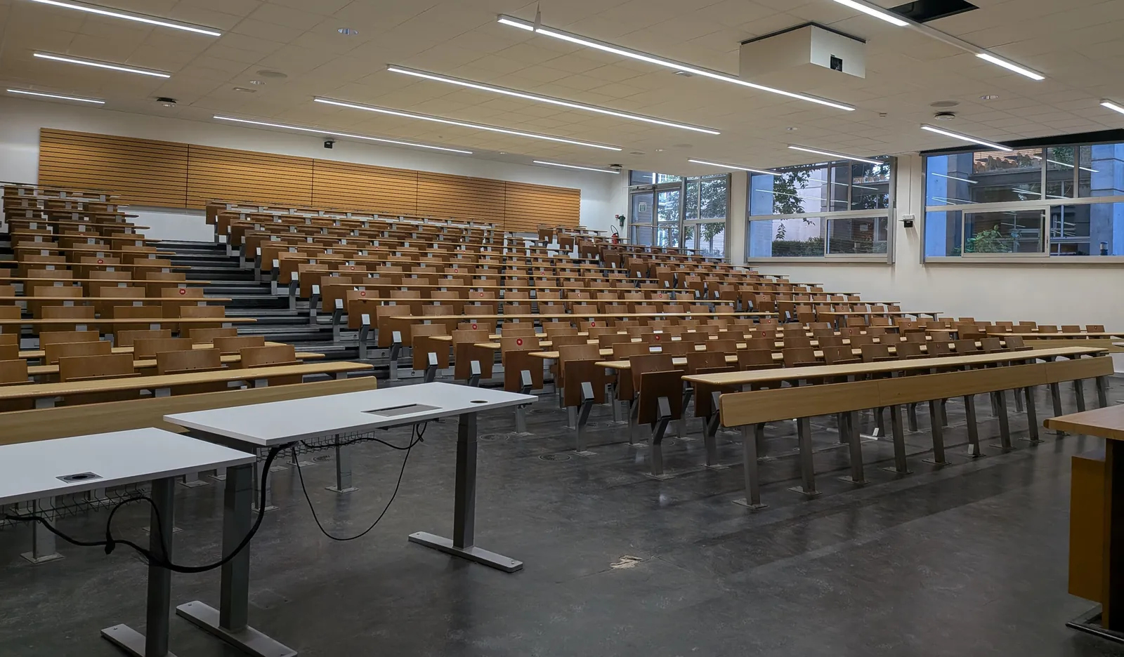 A large empty lecture theatre with light wooden seats, seen from the rostrum, with large windows on the right