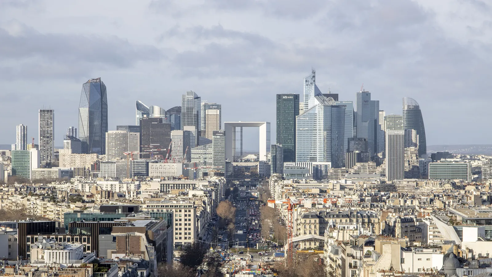 The office towers of La Défense and the Grande Arche, seen from a distance over the rooftops of Paris under a cloudy sky