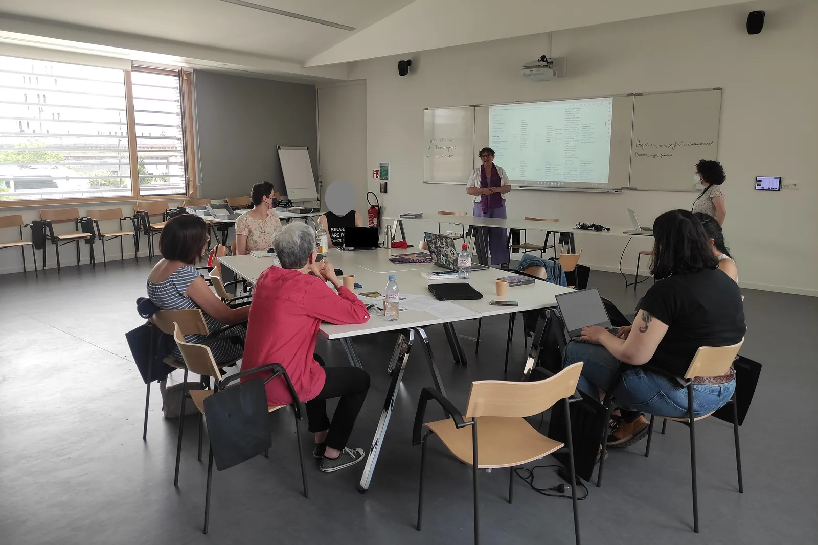 A wide shot of a training room: participants seated around tables with their laptops, a female trainer standing in front of a screen