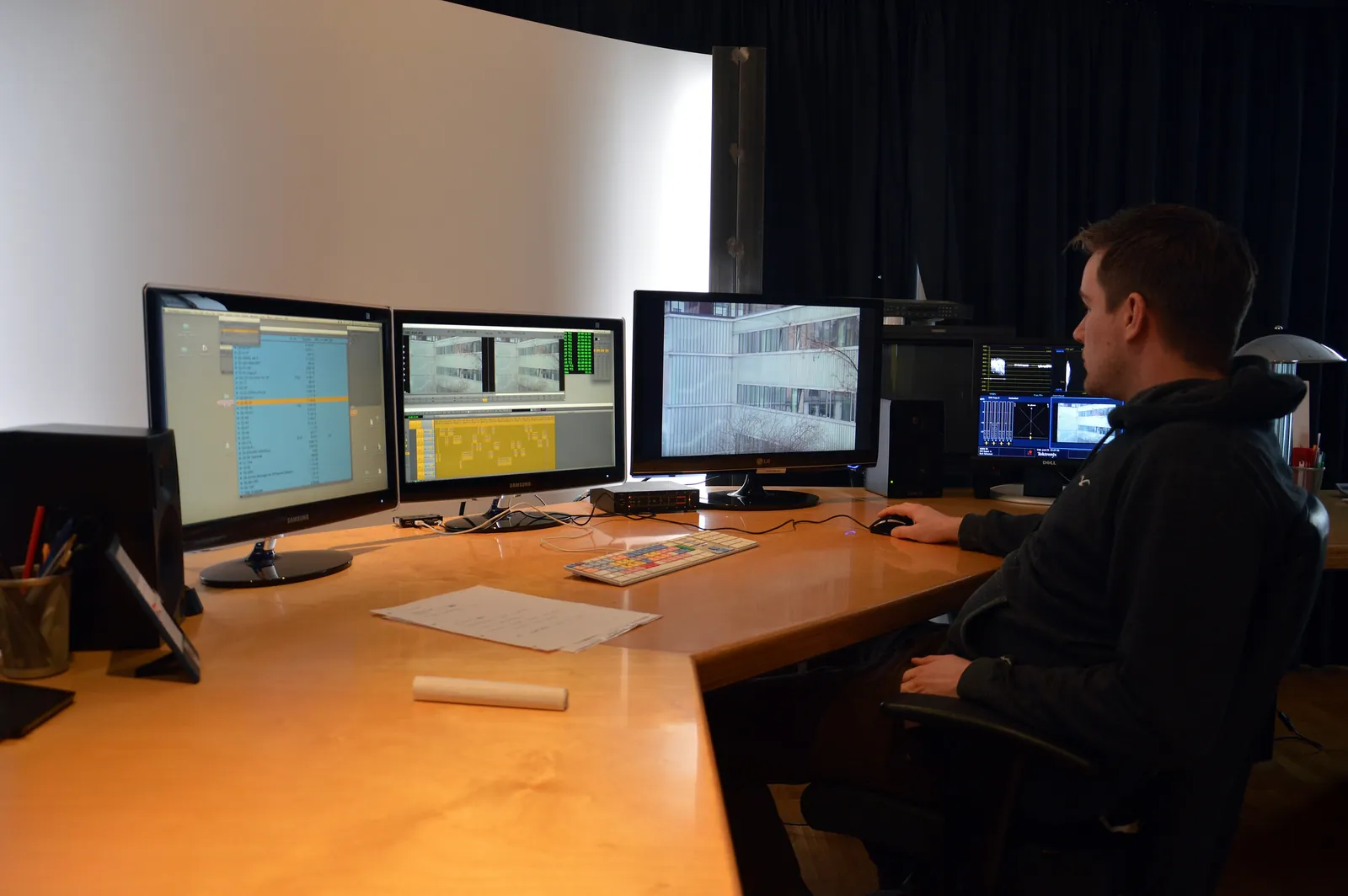 An editor seen in three-quarter rear view, in front of 3 screens, in a video editing suite with black curtains