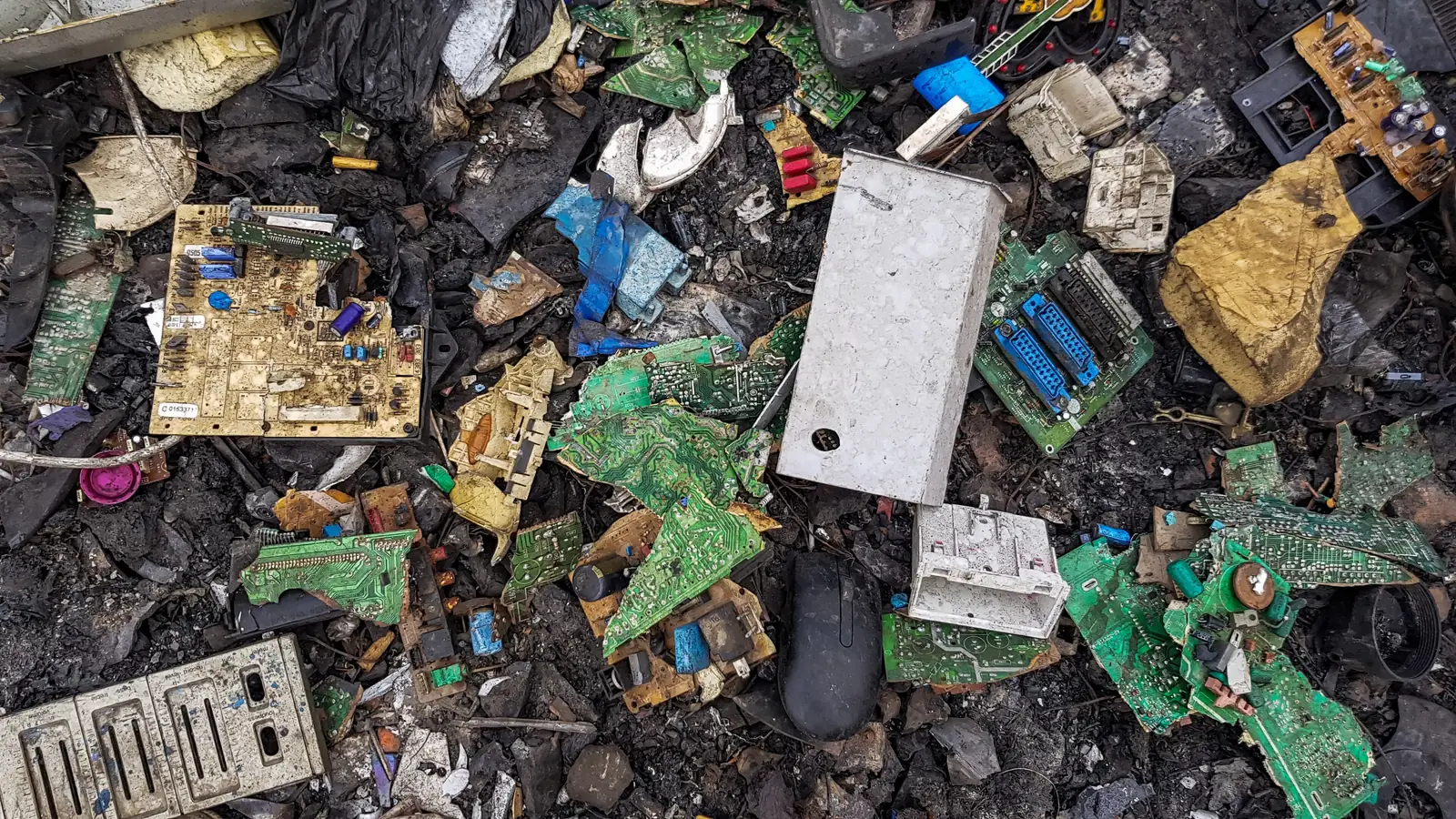 Broken circuit boards, casings and cables mixed together on blackened ground, in an open-air dump
