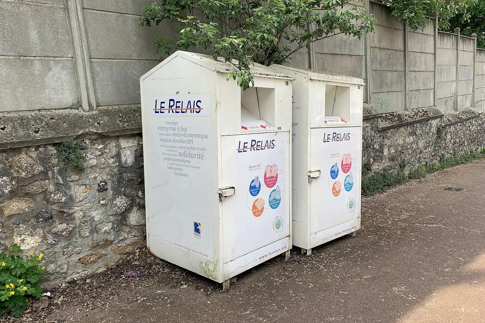 2 white Le Relais clothing collection containers on a pavement beside a stone wall