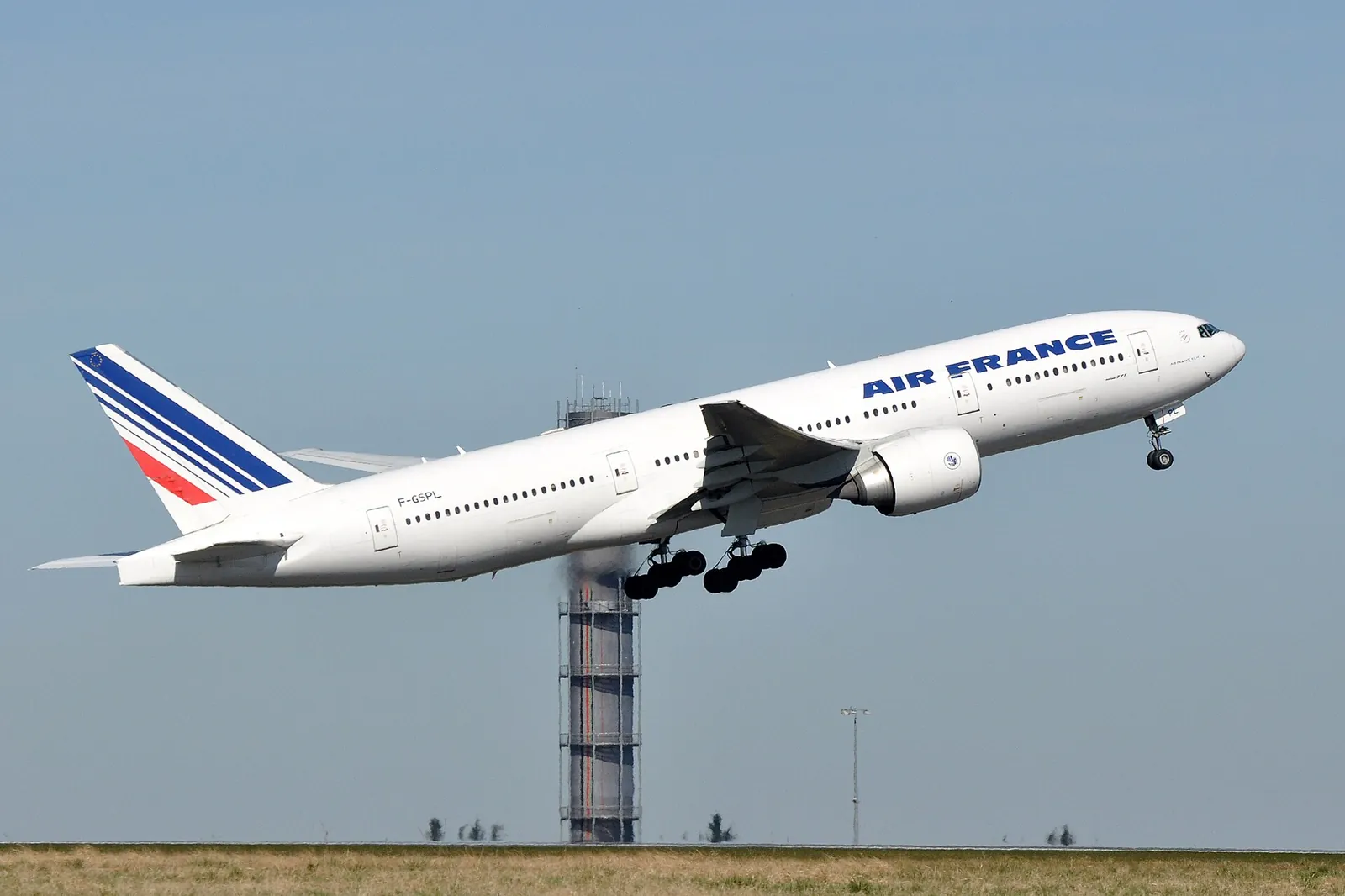 A long-haul airliner taking off, nose raised and landing gear still down, in front of a control tower