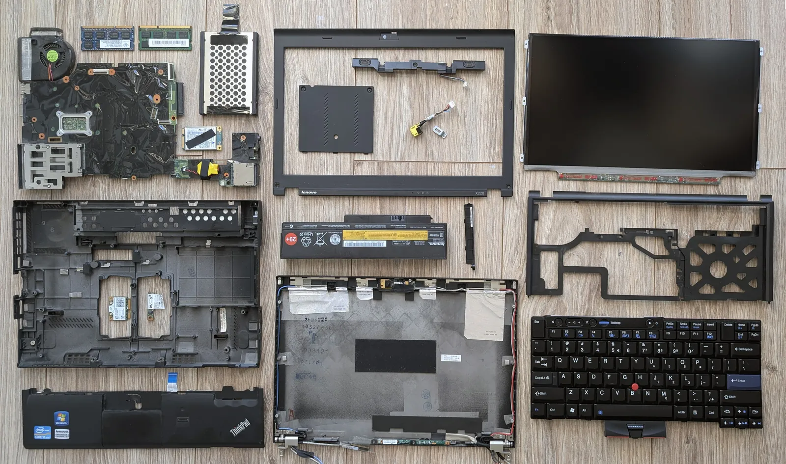 A laptop fully taken apart, its parts laid out on a wooden floor: motherboard, screen, battery, keyboard