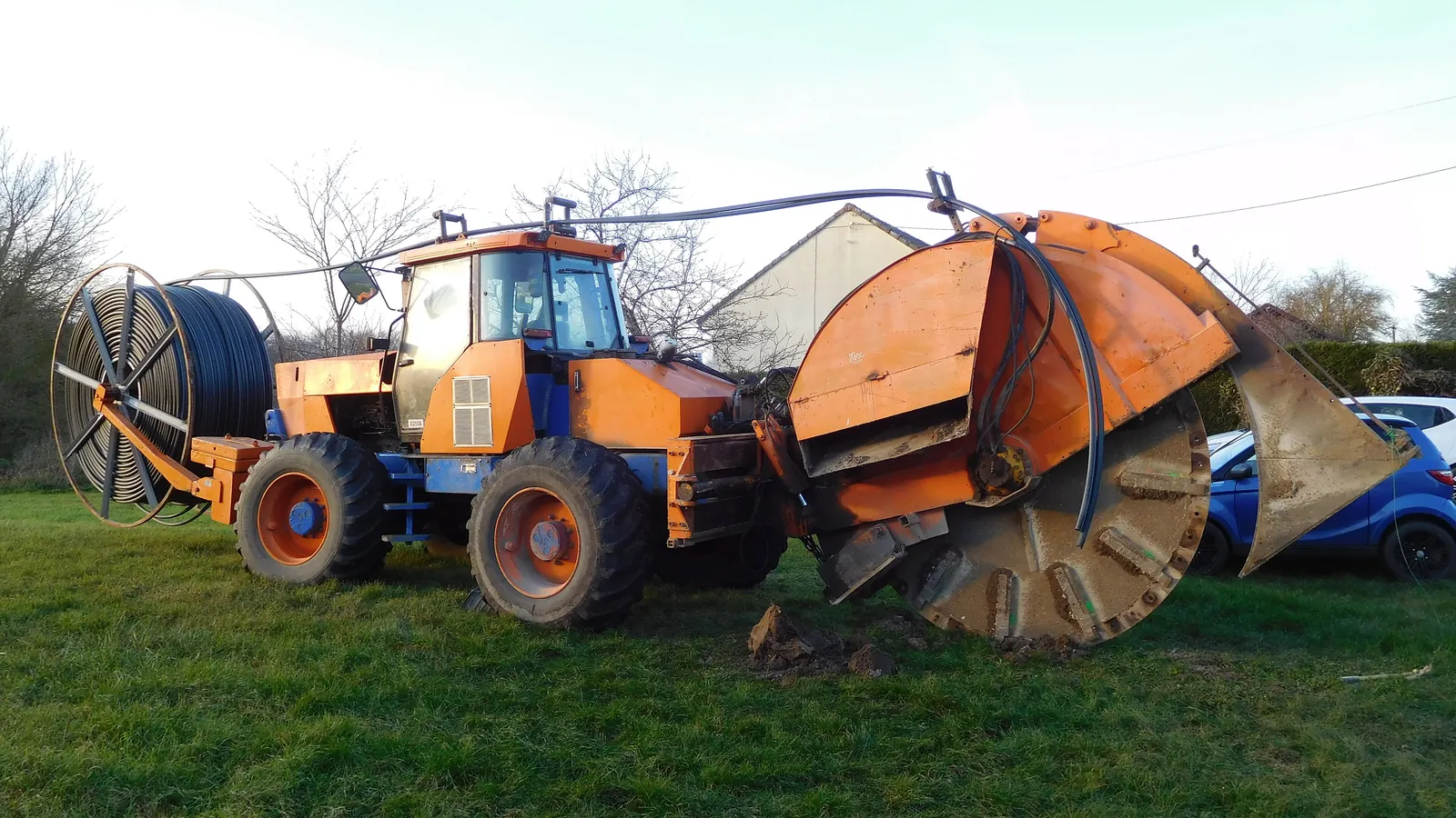 An orange construction machine unrolls a black duct into a trench at the edge of a meadow