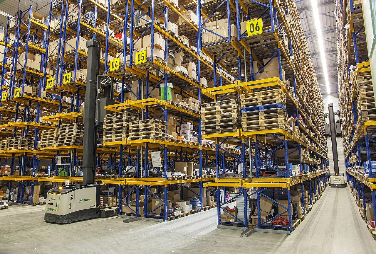 Aisle of a warehouse with high racking loaded with pallets and cardboard boxes, with a forklift truck