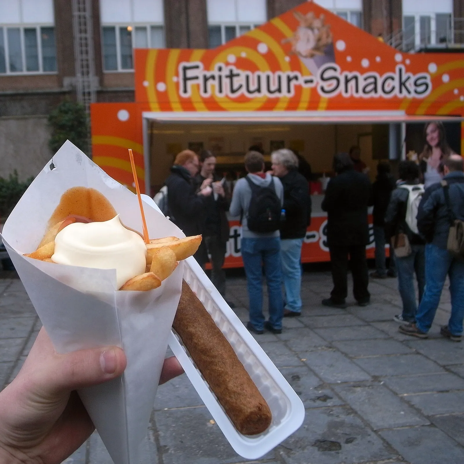Paper cone of chips with mayonnaise and a frikandel sausage, held in front of a chip shop