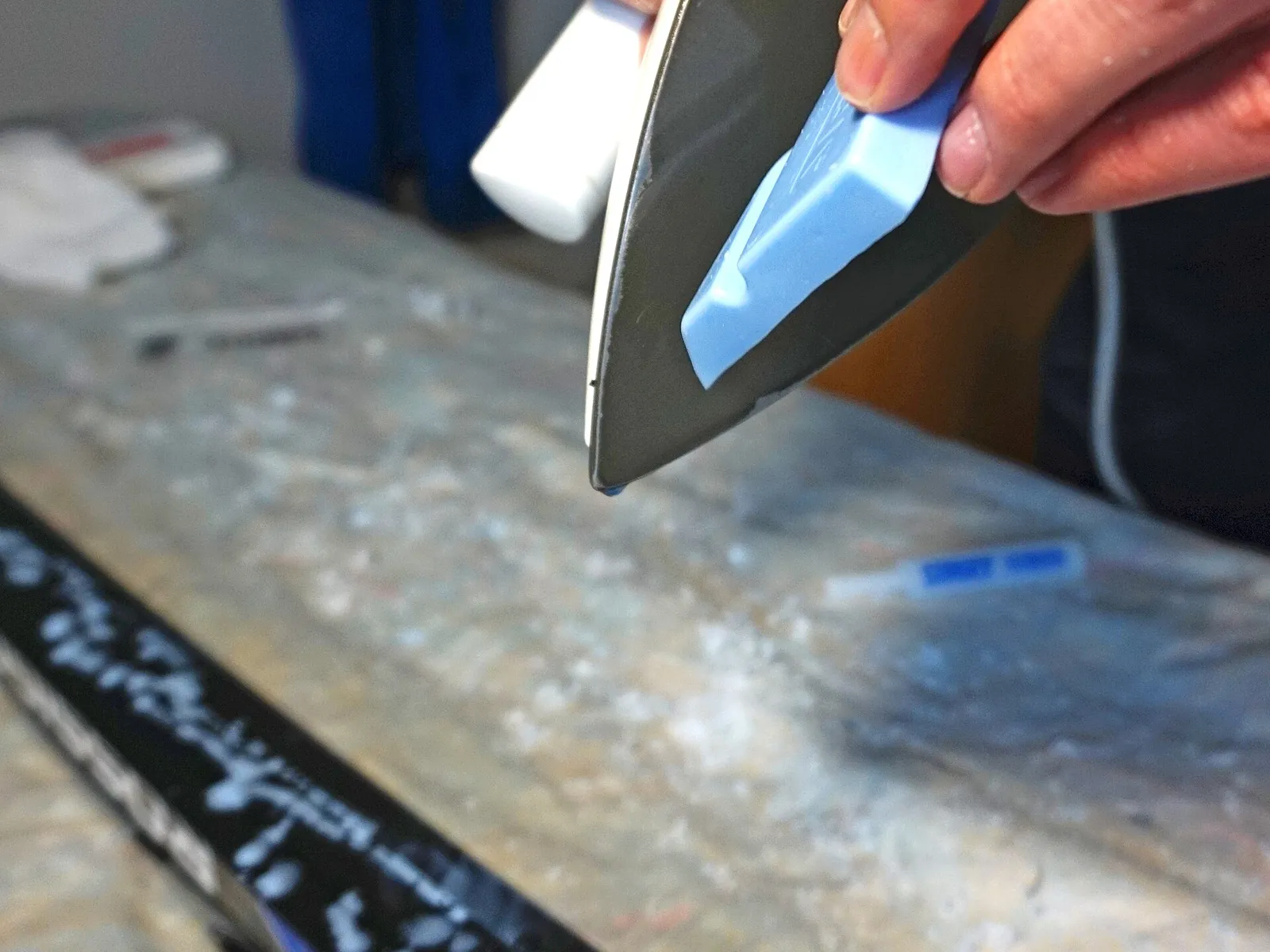 A waxing iron melting a blue bar of ski wax onto the black base of a ski resting on a workbench