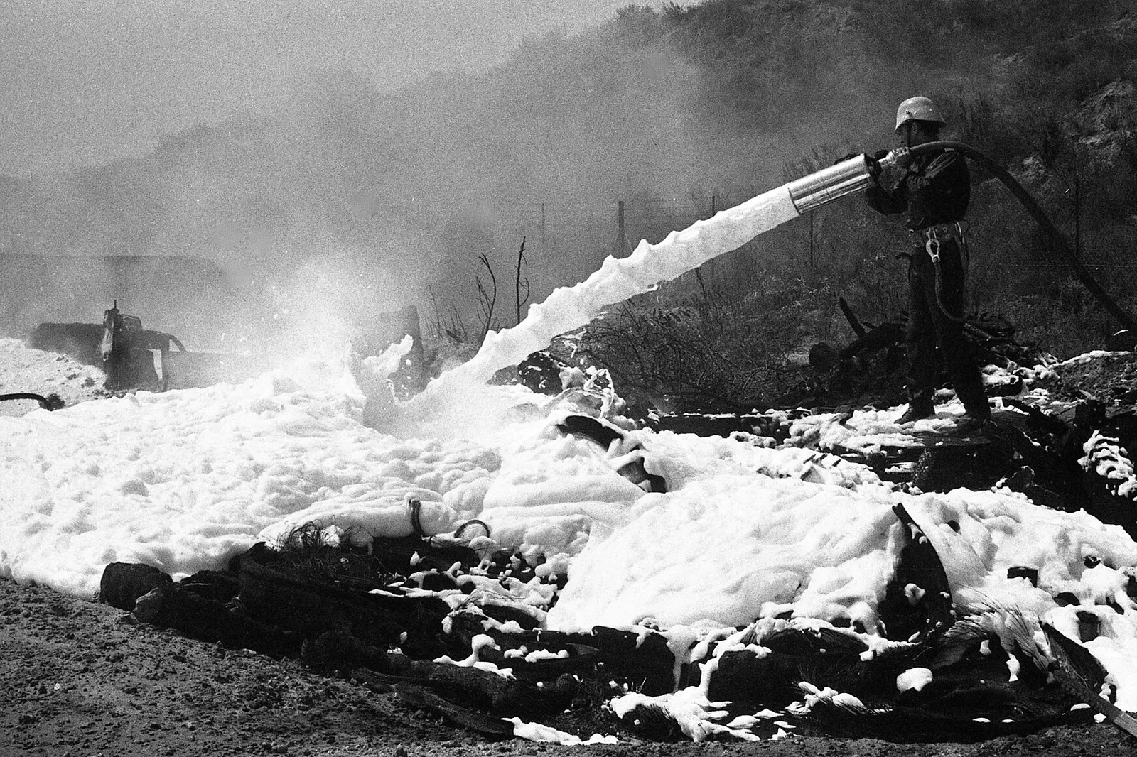 Black-and-white photo of a firefighter spraying foam onto a debris fire, which is covered by a white layer