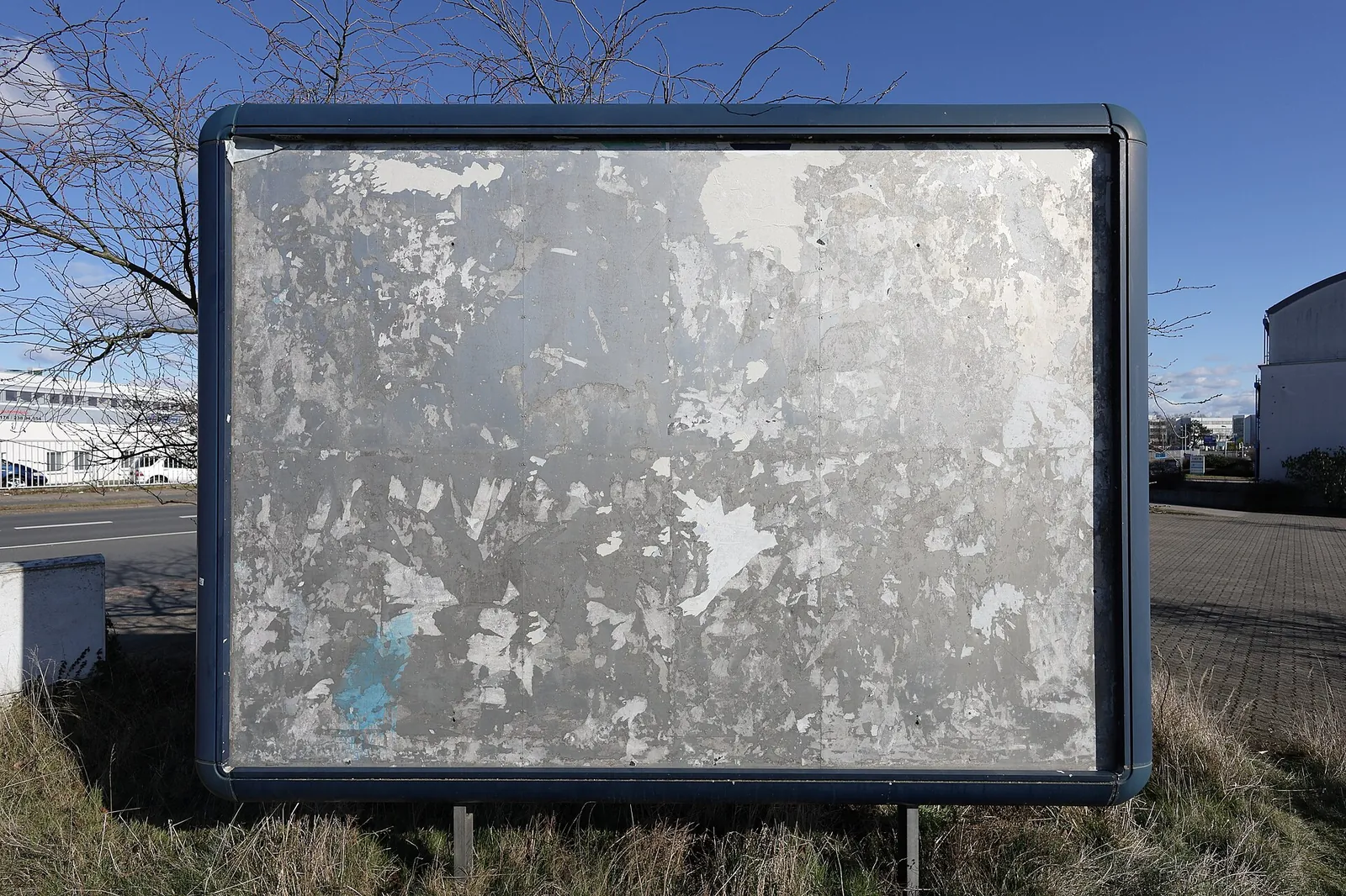 Empty billboard with torn-off posters beside a car park under a blue sky, leafless trees in the background