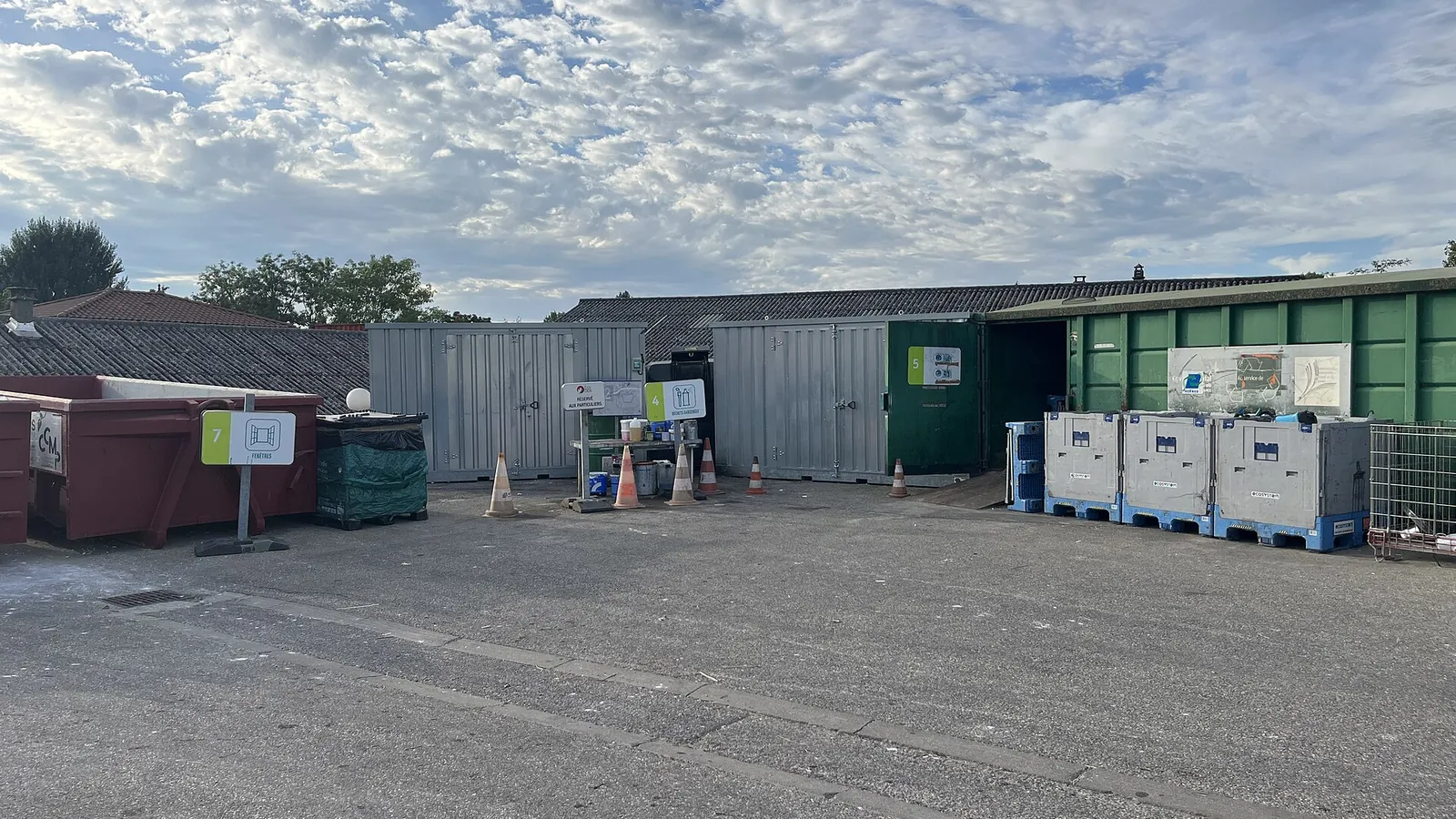 Drop-off area at the Miribel waste centre in Ain: a red skip marked by a "Fenêtres" sign, metal containers and pallet boxes lined up under a cloudy sky