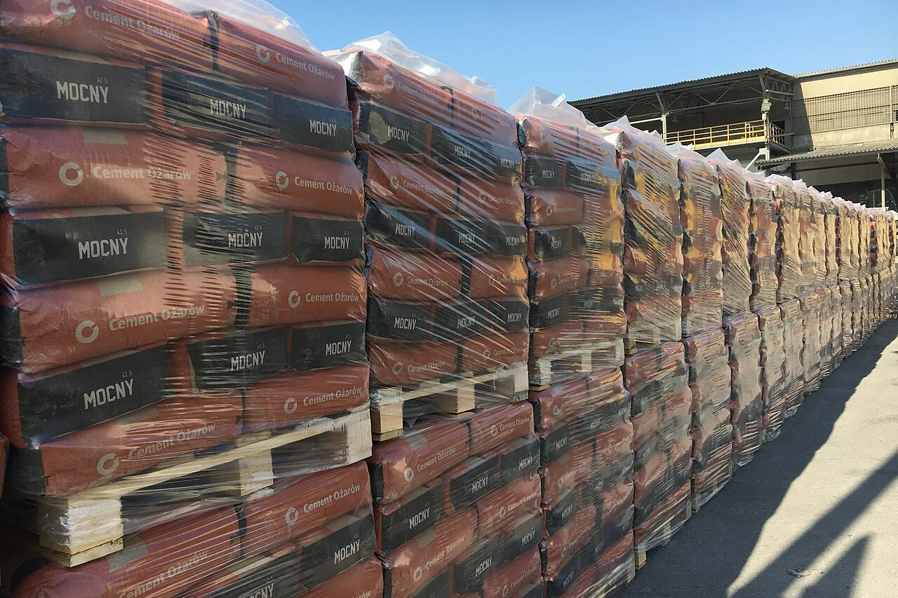 Long row of pallets of red and black cement bags, wrapped in clear stretch film, under a blue sky