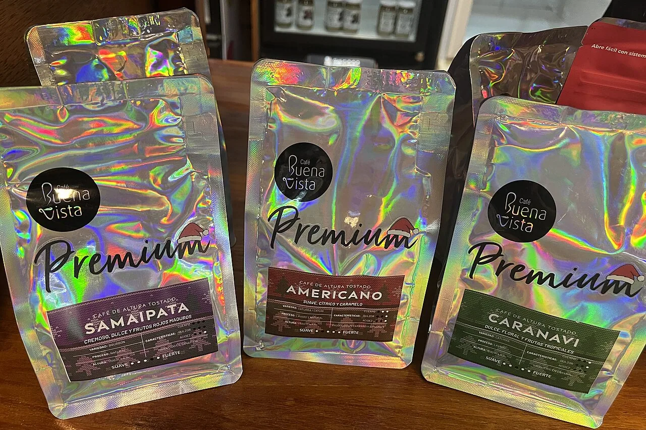 Three flat-bottomed coffee pouches in silver holographic film, standing upright on a wooden table