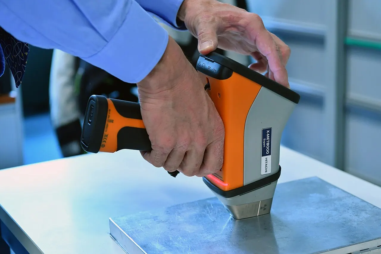 A technician places the head of an orange and black handheld X-ray fluorescence analyser on a metal plate lying on a table