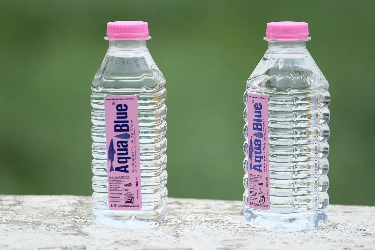 2 small bottles of clear PET water, with a pink cap and a pink and blue label, standing on a low wall in front of a green background