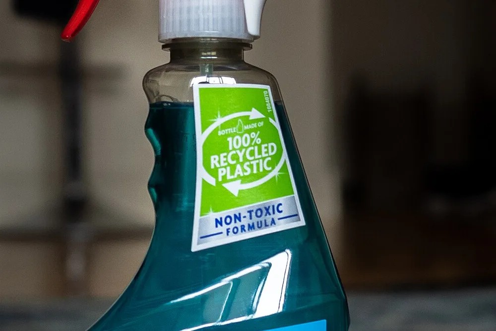 Windex glass cleaner spray bottle, with a green badge reading "Bottle made of 100% recycled plastic" on the neck