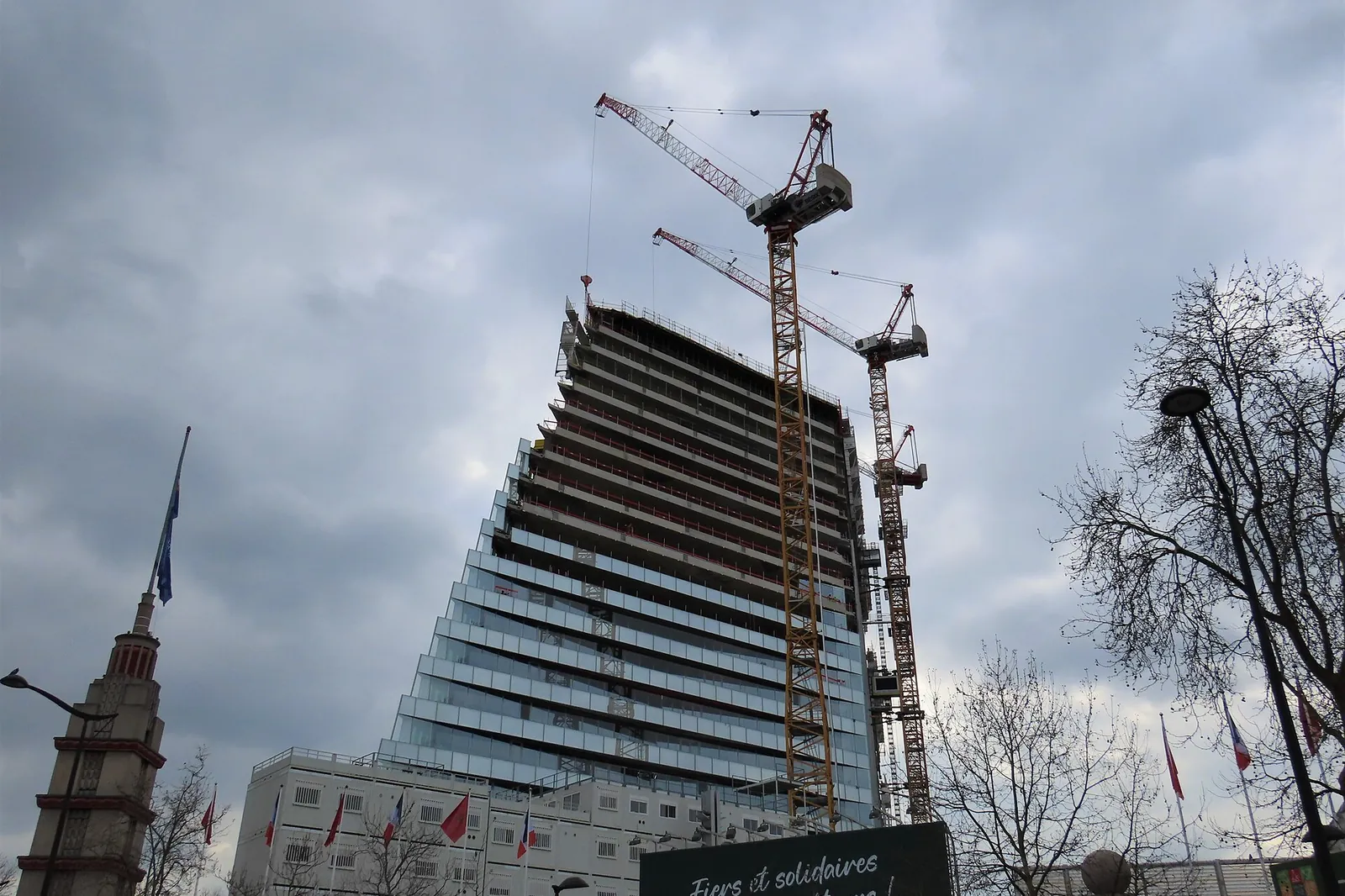 Glazed office tower under construction in Paris, surrounded by 2 cranes under a cloudy sky
