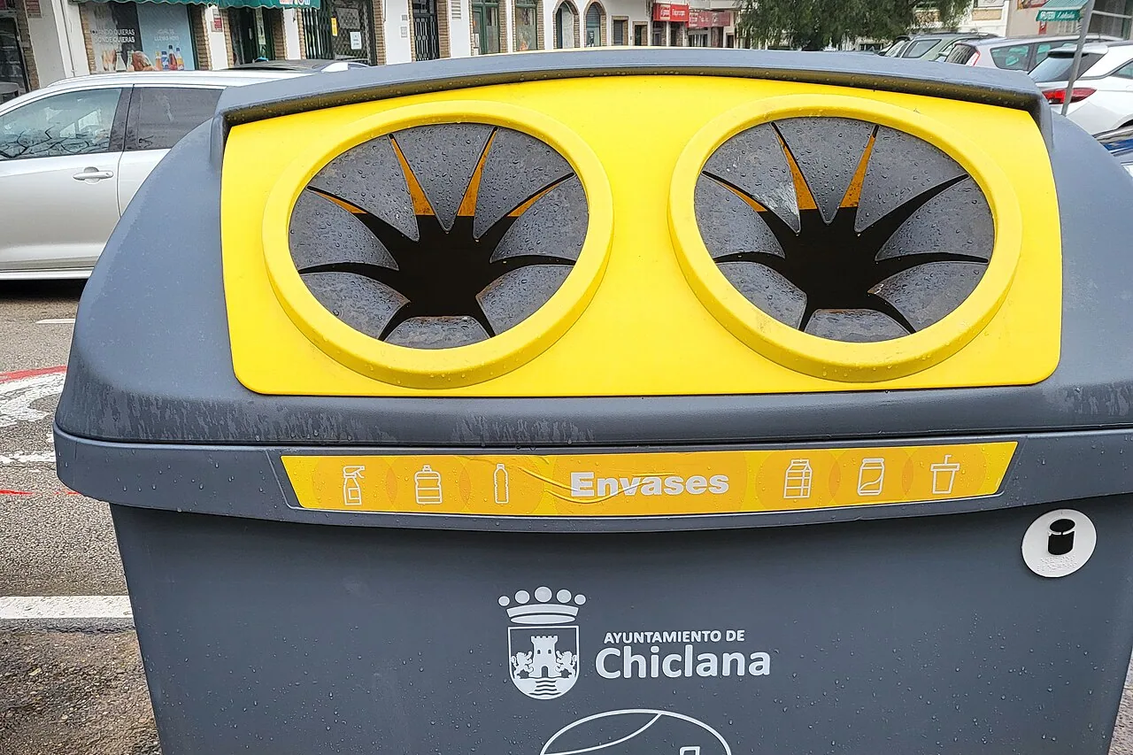 Grey container with a yellow lid pierced by 2 round openings, with the "Envases" banner and the coat of arms of the Chiclana town hall, at the edge of a street