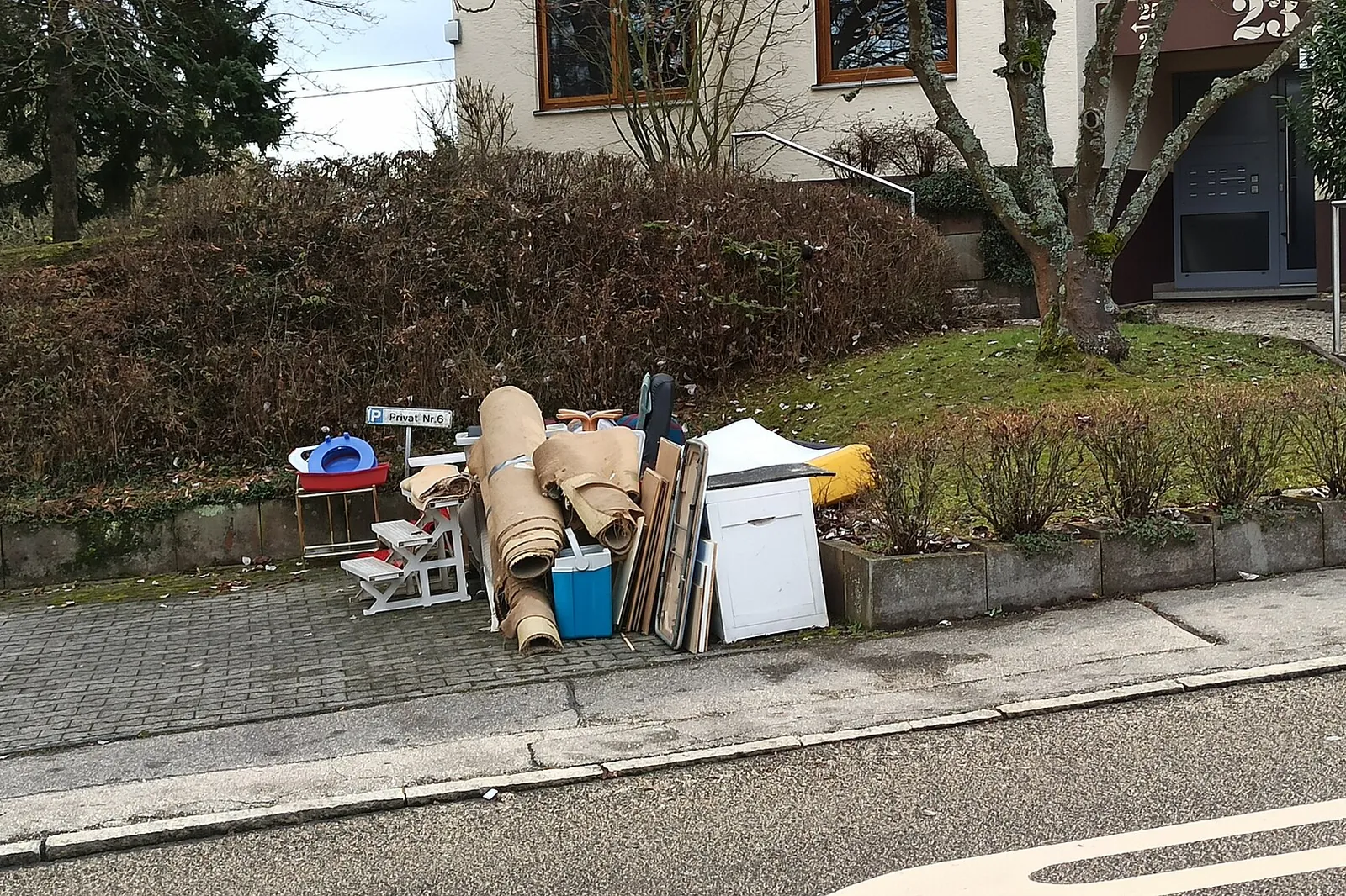 Bulky waste left at the edge of a car park in front of a house: rolls of flooring, a white piece of furniture, a stepladder, a cool box and wooden panels