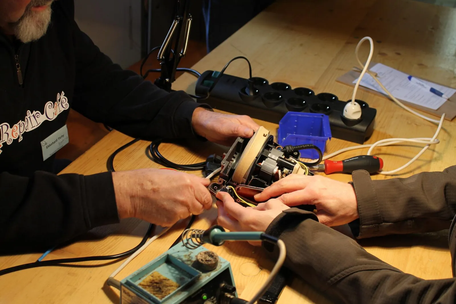 A volunteer and visitor repair the motor of a small household appliance on a wooden table beside a soldering iron