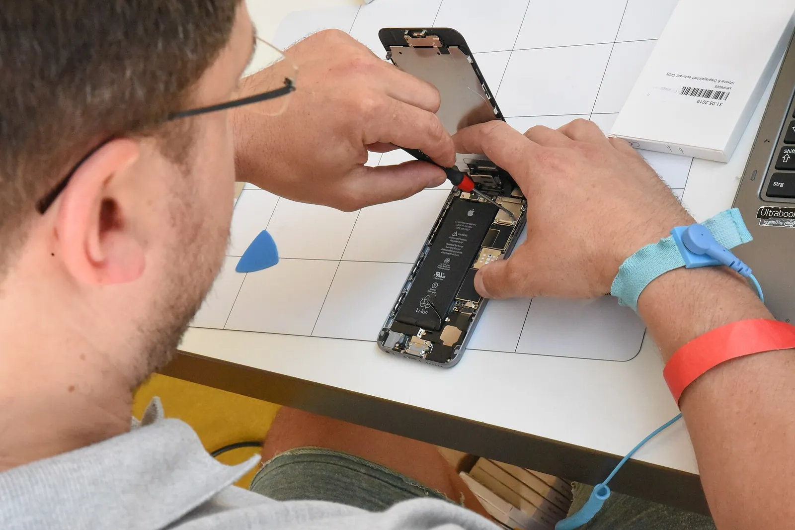 Hands opening a smartphone on a work mat, with the lifted screen exposing the battery, during a repair workshop
