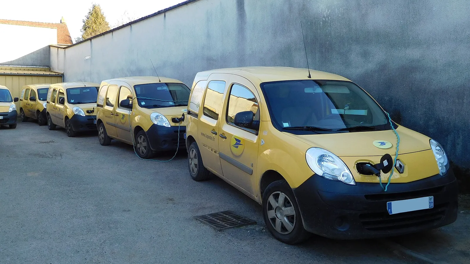 A row of yellow La Poste electric vans plugged into charging points along a wall